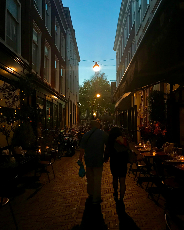 A couple walks hand-in-hand down a dimly lit alley flanked by buildings, with outdoor café seating and bicycles parked along the sides.