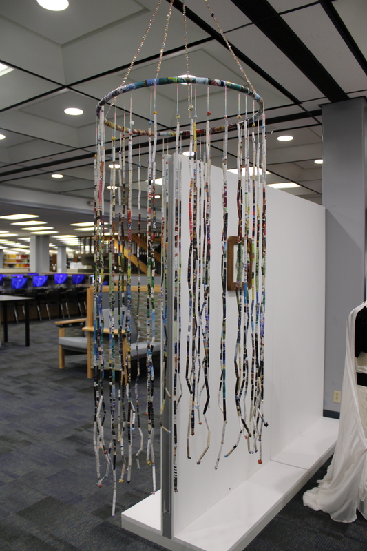 The installation consists of a circular frame from which numerous thin, colorful strips hang vertically. The strips appear to be made of tightly rolled paper, possibly from magazines or posters, and are suspended at various lengths, creating a curtain-like effect. T
