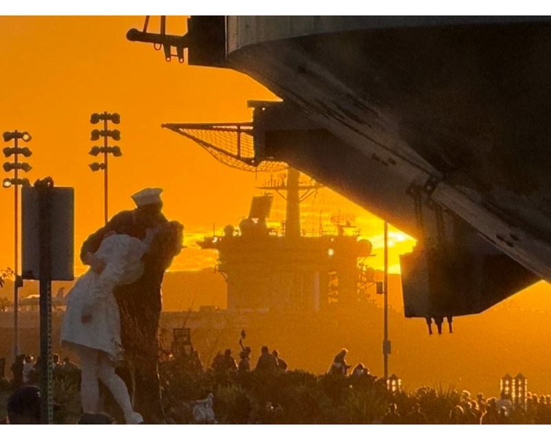 A statue of a sailor kissing a woman in a white dress is silhouetted against an orange sunset, with a ship and people in the background.