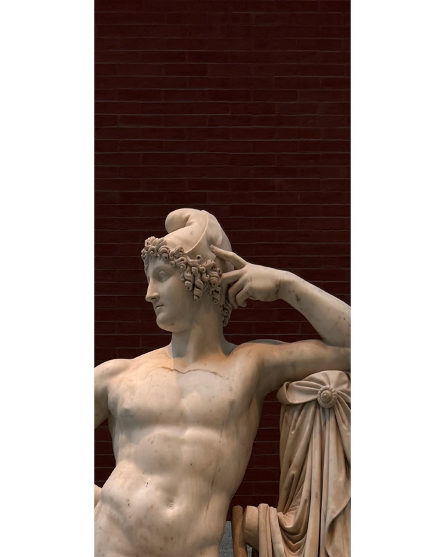 A marble statue of a muscular male figure with a laurel wreath, posed elegantly with one arm resting behind his head, set against a deep brick-red background.