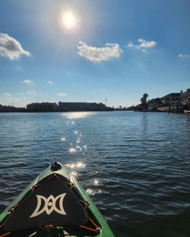 Front end of a green kayak extends into a calm body of water. The kayak has a black well cover, connected to the kayak with orange cording. Buildings and palm trees can be seen on the surrounding shoreline. The bright sun and a few clouds are in the sky. The sunlight sparkles on the water's surface.