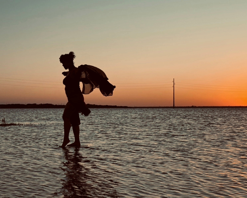 Silhouette of a woman wading in shallow water at sunset, holding a flowing fabric, with vibrant orange and blue sky in the background.