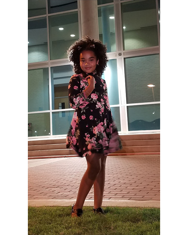 Color photograph of a young person in floral dress, posing in front of a building at night