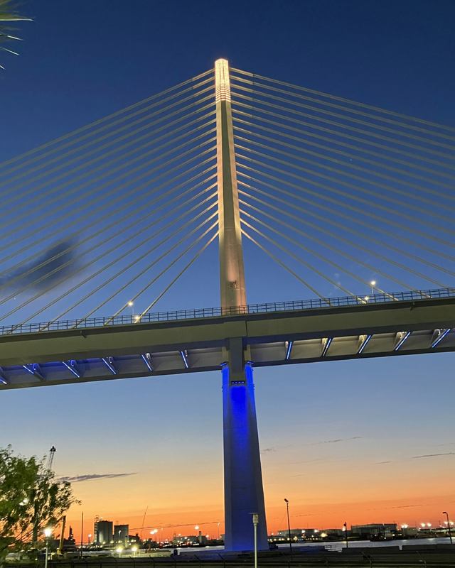 Suspension bridge at dusk with a lit support tower and blue lights, set against a gradient sky transitioning from blue to orange.
