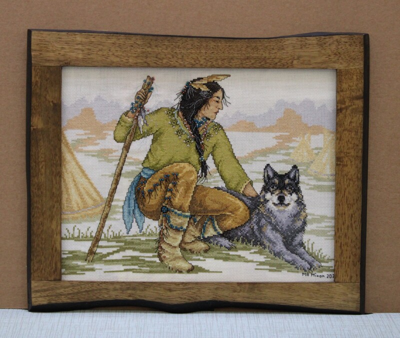 Framed cross stitch of a person with a staff sitting beside a wolf, set against a muted landscape.
