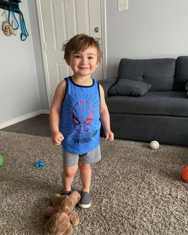 A young person in a sleeveless blue Spiderman t-shirt, shorts, and grey slip on shoes, stands in the middle of the room, smiling at the viewer. The figure is standing on a tan shaggy rug with toys strewn about the floor. The rug is in front of a dark grey couch along a wall with a closed door on the left.