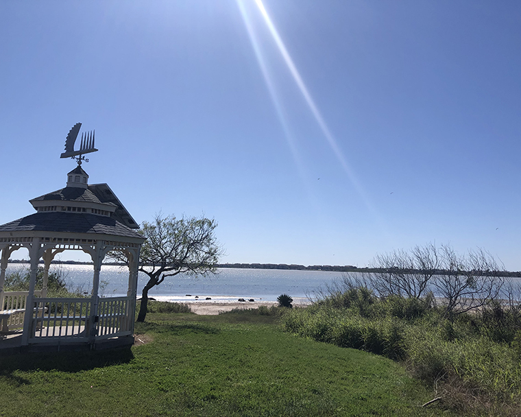 Color photograph of sunbeams in a landscape including a gazebo on a grassy shoreline 