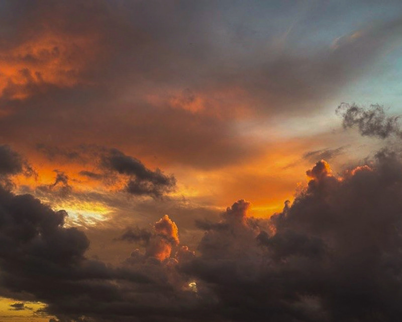 A clouded skyscape with orange, red, and yellow reflections. The clouds furthest from the viewer are soft and smooth, with the closer clouds having more structure and definition.