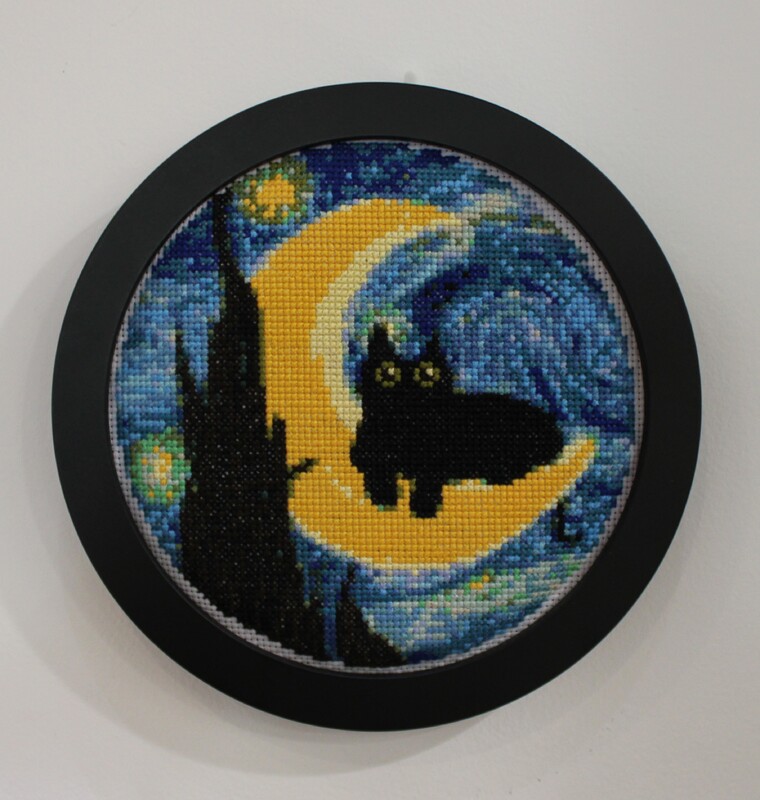 Embroidery hoop with black cat cross-stitch art with moon and star background.
