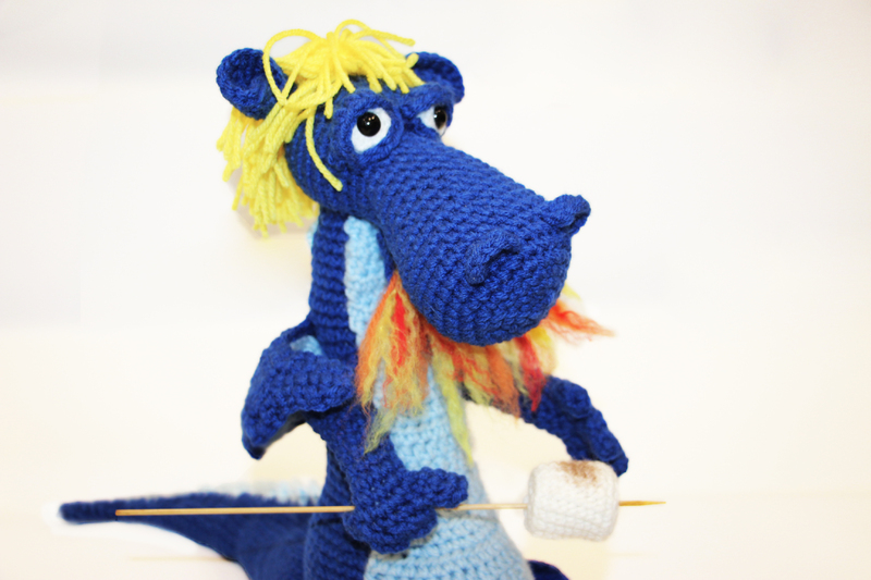 Close up view of crochet figure of a blue dragon. The dragon has yellow hair and is breathing fire to roast a marshmallow on a stick.