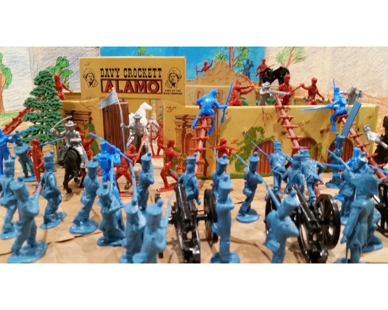 A colorful diorama of toy soldiers depicting a battle at the Alamo, featuring blue soldiers in formation, red soldiers climbing, and a cannon in the foreground.