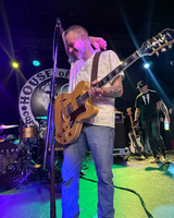 A guitarist in a patterned shirt plays on stage with colorful lighting, while a band member stands behind him. A drum set is visible in the background.
