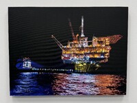 Offshore oil platform at night, illuminated with yellow and orange lights, with a spotlight from a nearby vessel reflecting on the ocean.

