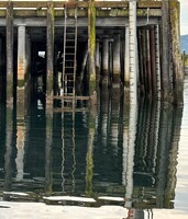 Artist: Kristina Alexander. 13x10” framed photograph titled Moody Waters 1. The photograph captures a pier-like structure rising out of a slightly rippled body of water. The structure is reflected on the water’s surface.