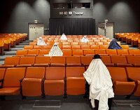 Artist: Alexis De León. A 12 by 15-inch archival pigment print, titled Sticking Around for the Stinger. The print captures the inside at a Theater with orange stadium seating. Six figures are scattered throughout the theater draped in fabric. Five with light fabric, one with dark covering. All wear hats and glasses on top of the draped covering. The perspective is from the front of the theater, looking at the audience.