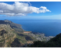 A panoramic view from a mountain showing rugged cliffs, a coastal town, and a deep blue ocean under a bright sky with scattered clouds.