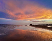 A picture is taken looking down the length of a beach shoreline. The viewpoint is from the shallow water's edge. The details of the shore, dunes, and structures are muted in the light. The sun is behind the horizon and creates vibrant colors on the clouds in the sky - the colors of the clouds are reflected on the water's surface.