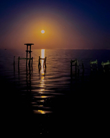 A bright moon rises over a calm water scene, with wooden posts silhouetted in the foreground and a warm glow illuminating the surface.