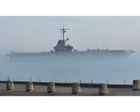 A large aircraft carrier partially obscured by fog, with concrete bollards in the foreground along the harbor's edge.