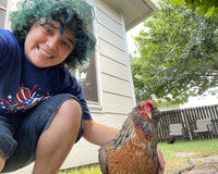 A smiling figure with blue green curly hair, crouches in the left side of the image, looking at the viewer. The figure is in a blue t-shirt with denim shorts, with their right hand embracing a brown chicken that is beside them on the ground. Both are in front of a light colored building, outside on a lawn, with paving stones and garden hose visible in the grass. The fence line, outdoor chairs, a second chicken, and tree canopy are in the background. 