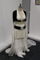 The image shows a dress form in an elaborate white and black outfit, displayed in a plain, brightly lit room. The outfit consists of a white, deep V-neck top with a black, short-sleeved bolero jacket layered on top. The jacket has a subtle floral texture. A wide black belt with a large circular silver buckle cinches the waist of the outfit. The pants are layered, featuring flowing, sheer white fabric that cascades to the floor, creating a train effect. Large circular embellishments adorn the skirt.