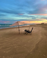 Two beach chairs are placed in the sand under a beach umbrella, next to a Reserved sign. The close shoreline is empty, with some walkers seen in the far distance. A few shorebirds are visible along the waters edge. Buildings can be seen in the distance. The sun is close to the horizon, reflecting peach and yellow hues onto the sky and clouds.
