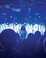 A live performer sings on stage in front of an audience, with a shimmering backdrop and blue lighting creating an atmospheric setting.