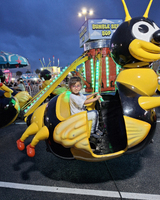 A child sits in a bumblebee-shaped carnival ride called "Bumble Bee Bop," surrounded by a festive atmosphere with colorful lights and rides.