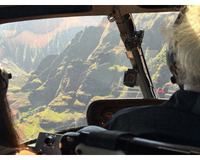 Interior view of a helicopter cockpit with two people wearing headsets, overlooking steep green mountains below.