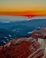 Sunset over a mountainous landscape with layers of red and brown hills, dotted with green trees, beneath an orange and blue sky.