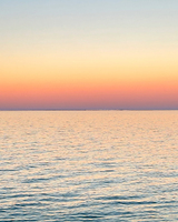 A calm seascape at twilight, displaying a smooth water surface reflecting soft hues of pink and blue, with a clear horizon.