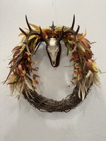 Artist: Diana Ivy. 36 by 30-inch mixed media titled Euro Mount Fall Wreath. A large grapevine wreath decorated with fall leaves and feathers hangs from a mounted animal skull with antlers.