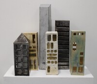 A group of ceramic sculptures resembling a city skyline on a white shelf, featuring simplified skyscraper forms in various colors and textures.
