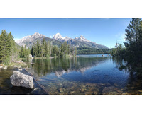 A clear, still body of water fills the foreground. Trees and snow covered mountains are in the background, topped with a lightly clouded blue sky. A large rock sits in the front left of the frame. Logs and stones can be seen under the water's surface.