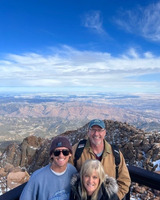 Three smiling figures pose for the camera in a corner railing, overlooking a rocky terrain. The figures are in cold weather clothing, two have on hats. The lightly clouded blue sky can be seen in the background. The picture appears to be taken from a great height and the horizon line is very far in the distance.