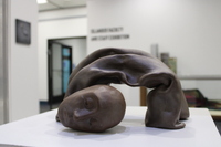 The image shows a sculpture placed on a white pedestal inside a gallery space. The sculpture is crafted from a material resembling bronze or dark clay, depicting a human figure in an arched position.

