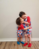 Two young people embrace in front of a tan wall with baseboard and wooden flooring visible. The two figures are wearing matching red, white and blue overall outfits with characters from Bluey (a television cartoon). Both have red t-shirts, the shorter person has blue socks, the taller in red socks.