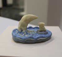 The image shows a small sculptural piece with a distinct aquatic theme. A prominent cream-colored shape, similar to a cresting wave or moon, rises from the blue base, surrounded by small, rounded white pieces that could represent foam or bubbles. Adjacent to the wave, there is a small mound with another cream-colored form atop it, possibly suggesting a rabbit on a rock or island. 
