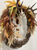 Artist: Diana Ivy. 36 by 30-inch mixed media titled Euro Mount Fall Wreath. A large grapevine wreath decorated with fall leaves and feathers hangs from a mounted animal skull with antlers.