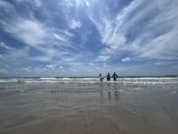 Three people with surfboards walking towards the ocean on a beach under a blue sky filled with wispy clouds.
