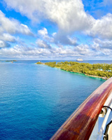 Bright blue water, vegetation, and other watercraft are seen from the deck of a ship. The wooden railing and the shadow of the ship can be seen in the lower right of the frame. The sky is bright with scattered clouds.