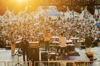 Image taken at a concert outdoors at sunset. The view is from behind the stage, the crowd is visible through the back of the band members and their equipment.