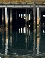 Artist: Kristina Alexander. 13x10” framed photograph titled Moody Waters 2. The photograph captures a pier-like structure rising out of a slightly rippled body of water. The structure is reflected on the water’s surface.