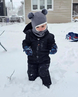 A child in a black snowsuit and gray pom-pom hat plays in deep snow outside a beige house.