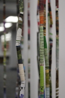 The installation consists of a circular frame from which numerous thin, colorful strips hang vertically. The strips appear to be made of tightly rolled paper, possibly from magazines or posters, and are suspended at various lengths, creating a curtain-like effect. T
