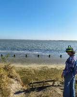 A woman in a colorful hat and shirt stands by a calm body of water with wooden posts visible in the foreground and a clear blue sky above.