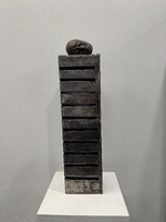 Artist: JinSun Kim. 24 by 7-inch sculpture of metal and clay. An upright tower of metal with nine slits on the left side of the piece. A sculpted head rests on top of the tower.