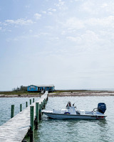 A boat with an outboard motor is tied to a wooden pier in a body of water. The pier leads to a distant shore in the background, leading away from the viewer. On the shore is a blue and white building with shrubbery and low grass. A blue sky with light clouds is visible in the background.