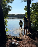 Two individuals by a tranquil lake, one wading in the water and the other standing on the shore, surrounded by greenery and a pair of flip-flops on the ground.