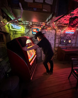 A figure with a brimmed hat and dressed in dark clothing leans against a juke box in a dark room. The stone walls are decorated with three neon signs advertising a variety of beer brands. The ceiling, trim, and doors are covered in dollar bills. The corner of a chair and a piano can be seen in the dim room. The space is lit by the light of the juke box, the neon signs, and strands of multi colored twinkly lights draped from the ceiling.